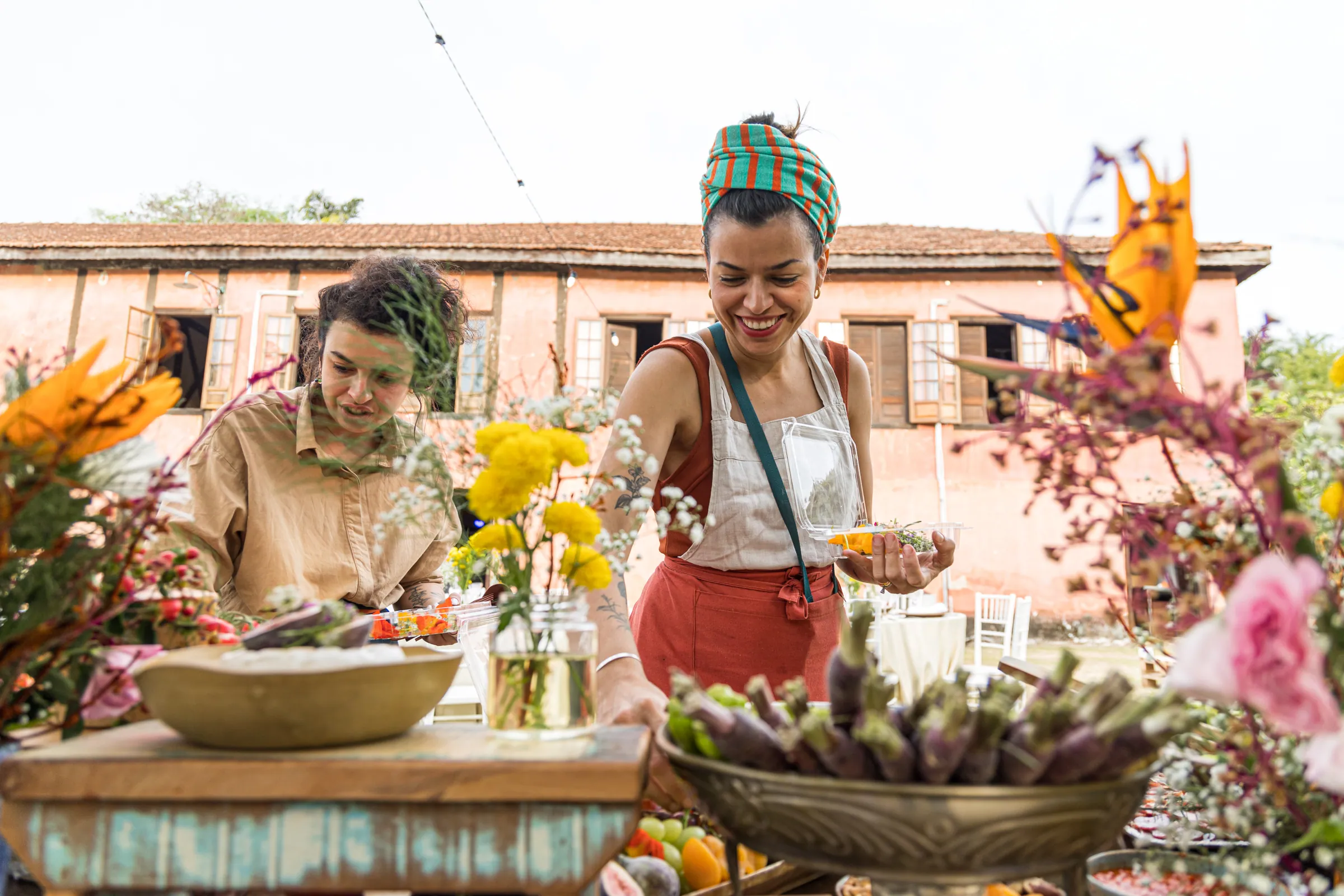 Duas mulheres arrumando a mesa de frutas e flores da festa, casarão rosa da Fazenda Cósmica ao fundo
