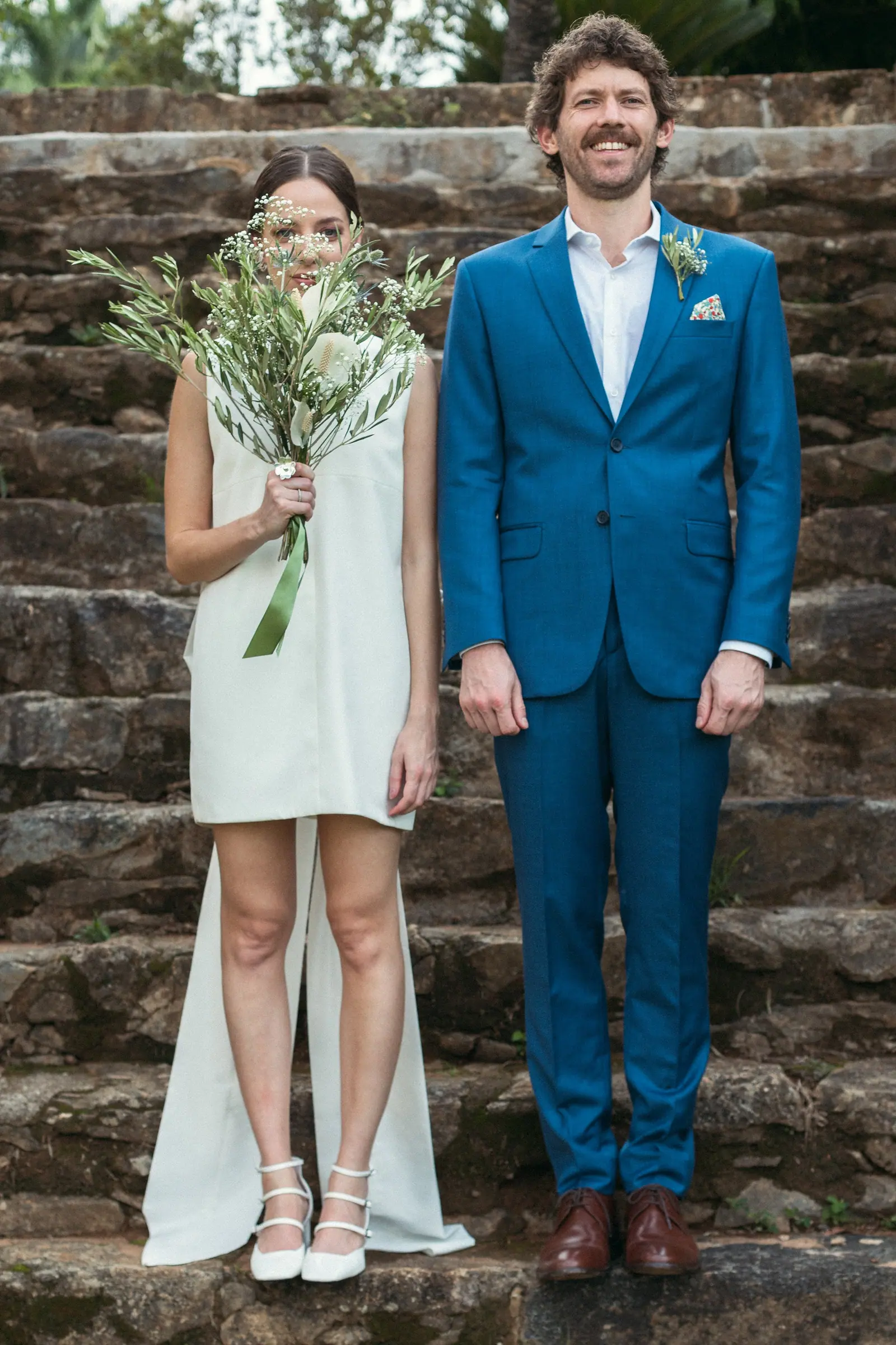 Retrato do casal recém-casado em uma escadaria de pedra, ela de vestido curto e buquê de flores silvestres, ele de terno azul