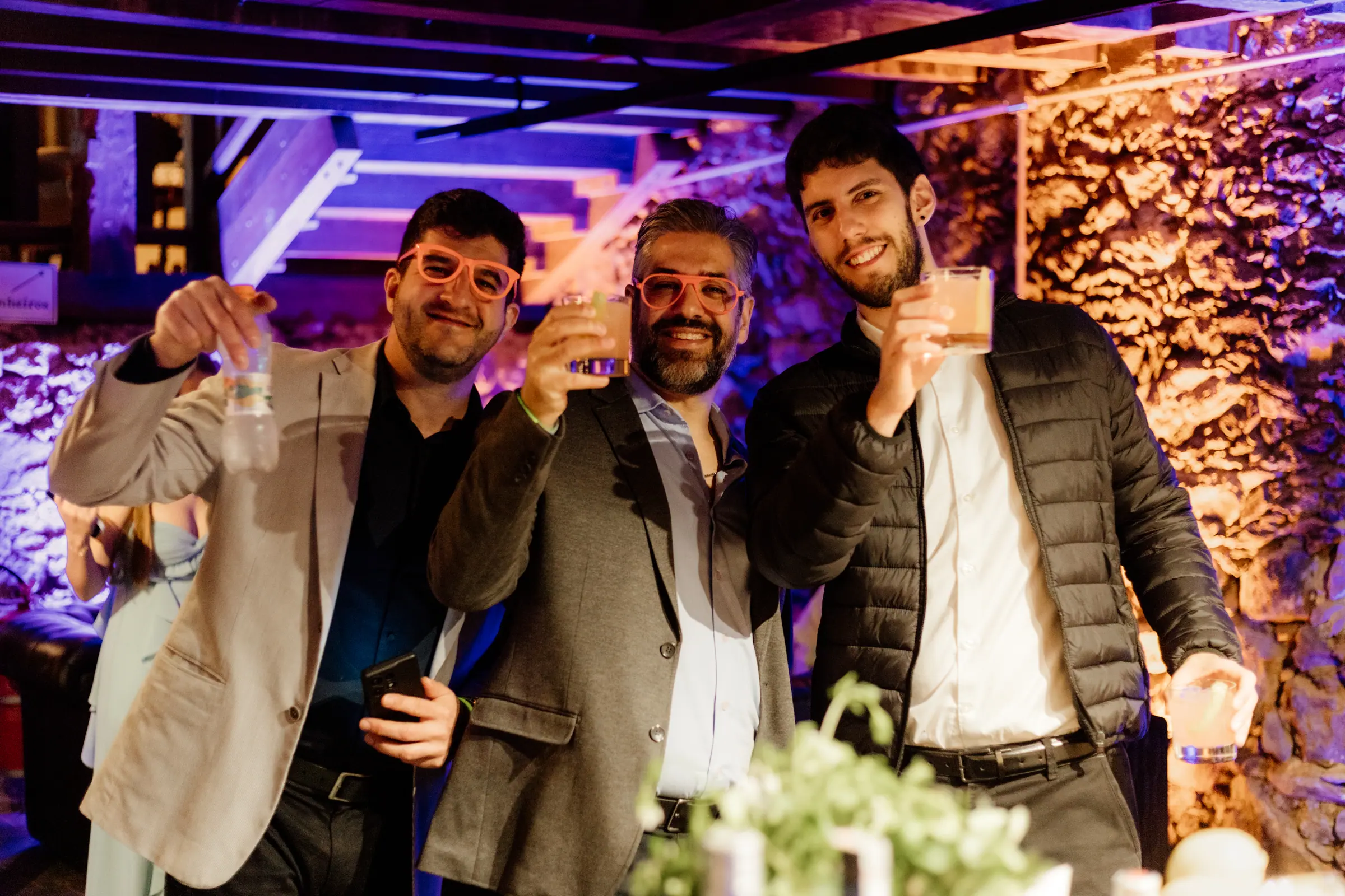 Três amigos brindando com drinques na festa, óculos de festa coloridos, iluminação roxa e dourada