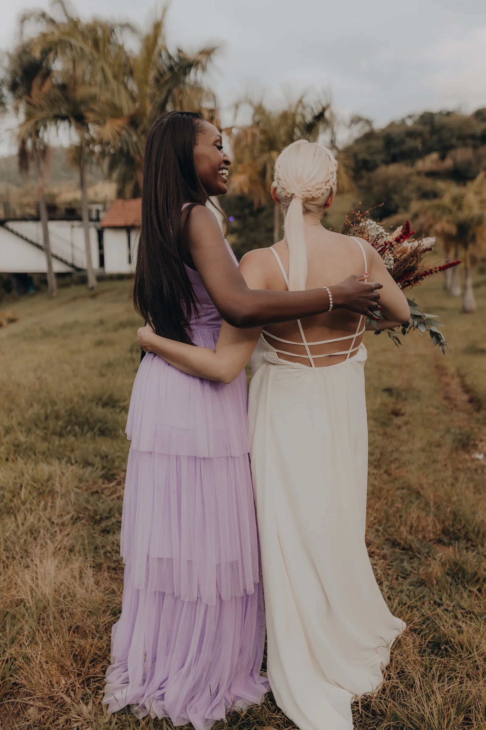 Convidada de vestido lilás abraçando a noiva, com buquê de flores secas e o campo da Fazenda Cósmica ao fundo