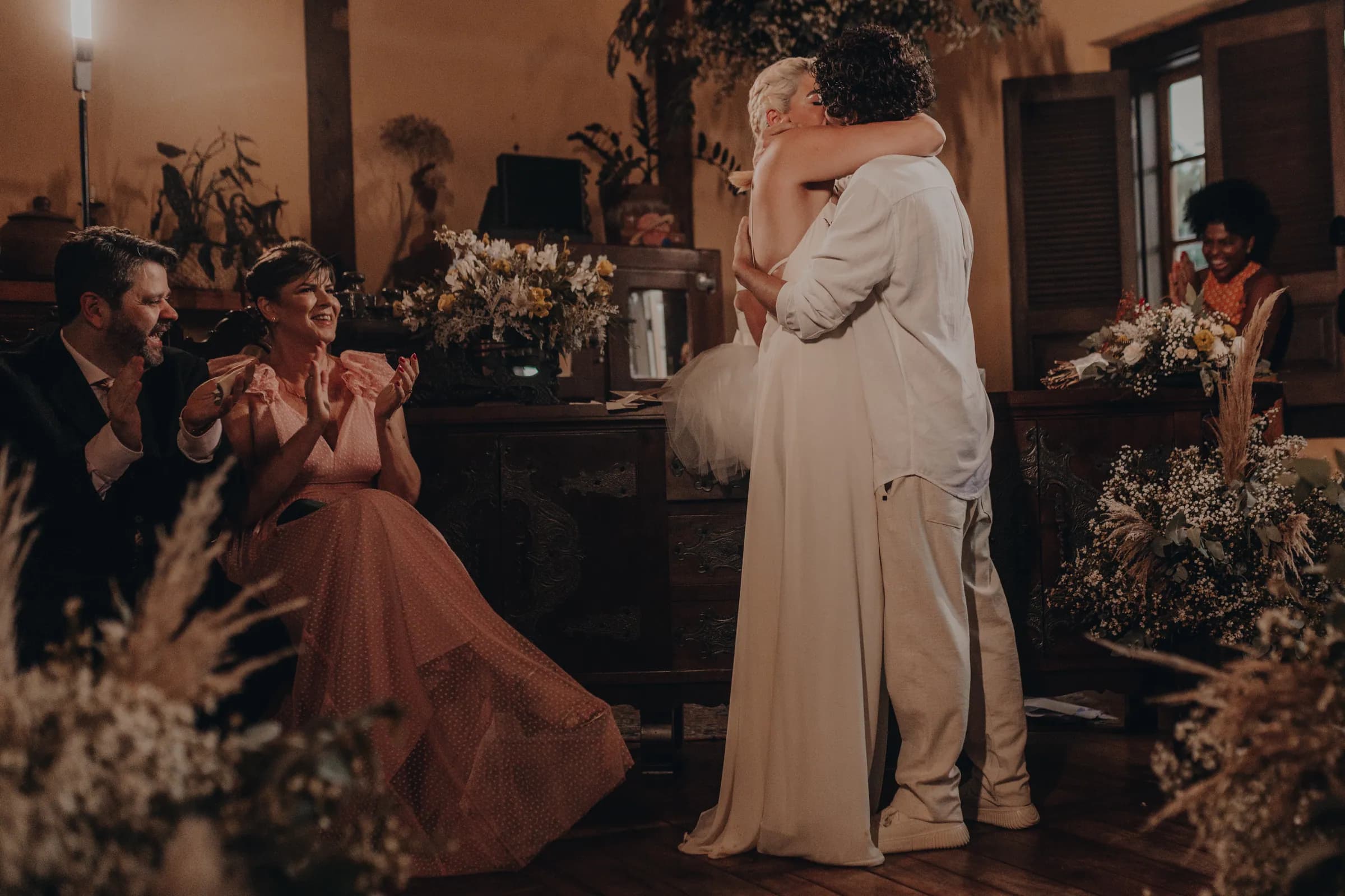 Abraço do casal no altar logo depois do sim, com madrinha aplaudindo em primeiro plano e arranjos de flor seca em volta
