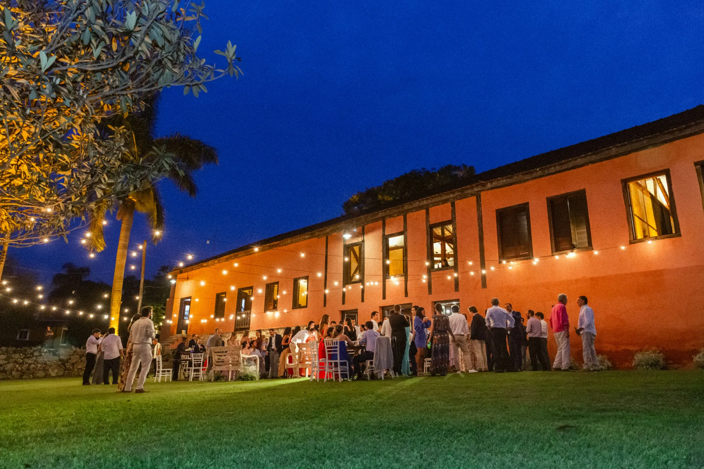 Festa de casamento à noite em frente ao casarão da Fazenda Cósmica iluminado por varal de luzes, convidados reunidos na grama
