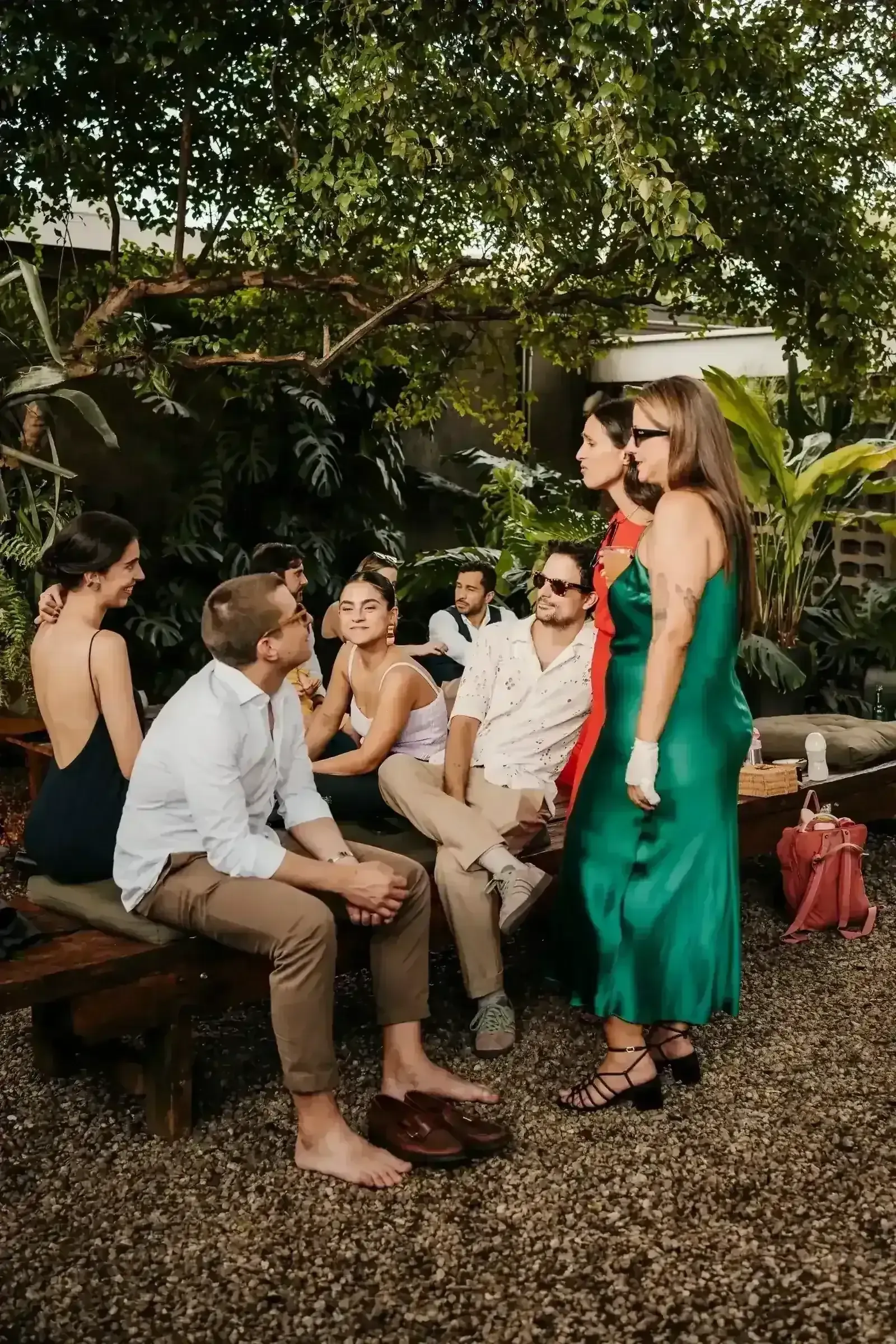 Convidados sentados no banco do jardim conversando ao fim do dia, com convidada de vestido verde em primeiro plano, em casamento na Fazenda Cósmica