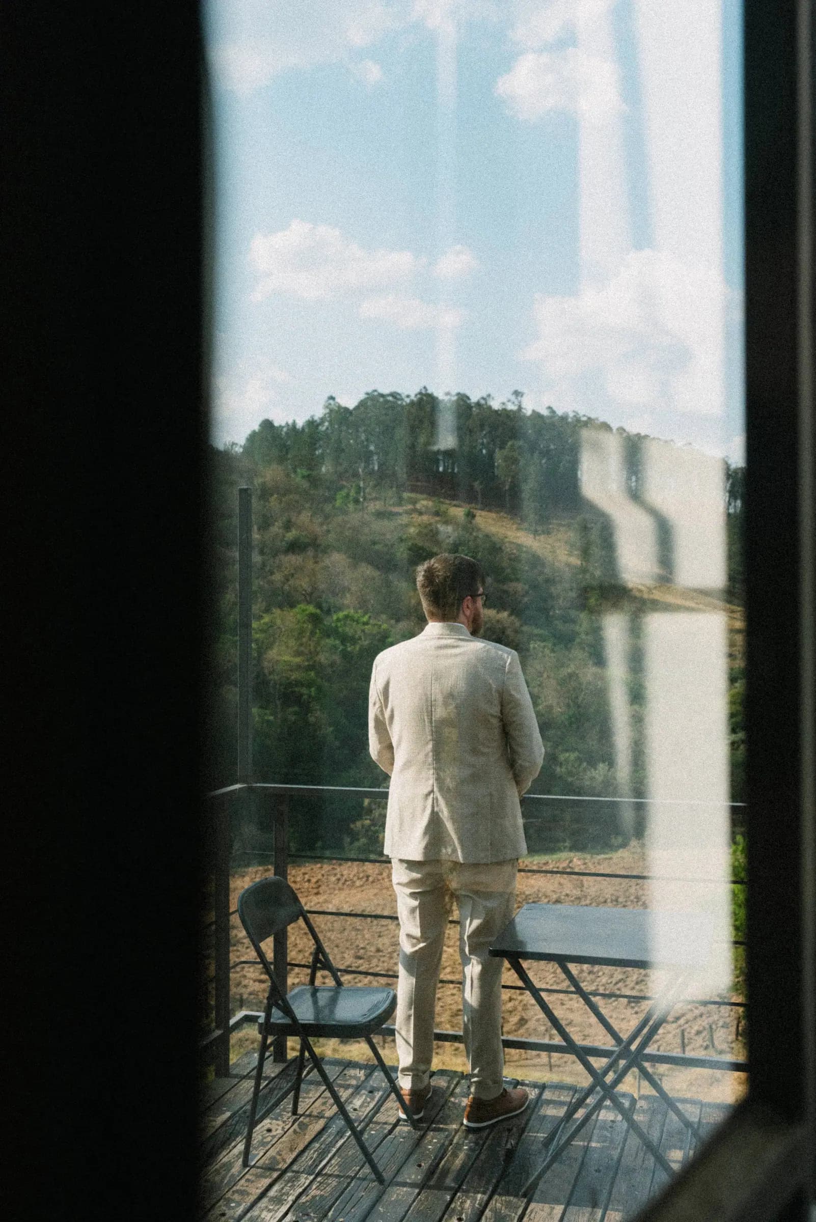 Noivo de costas na varanda da Fazenda Cósmica, observando o vale e a mata antes da cerimônia