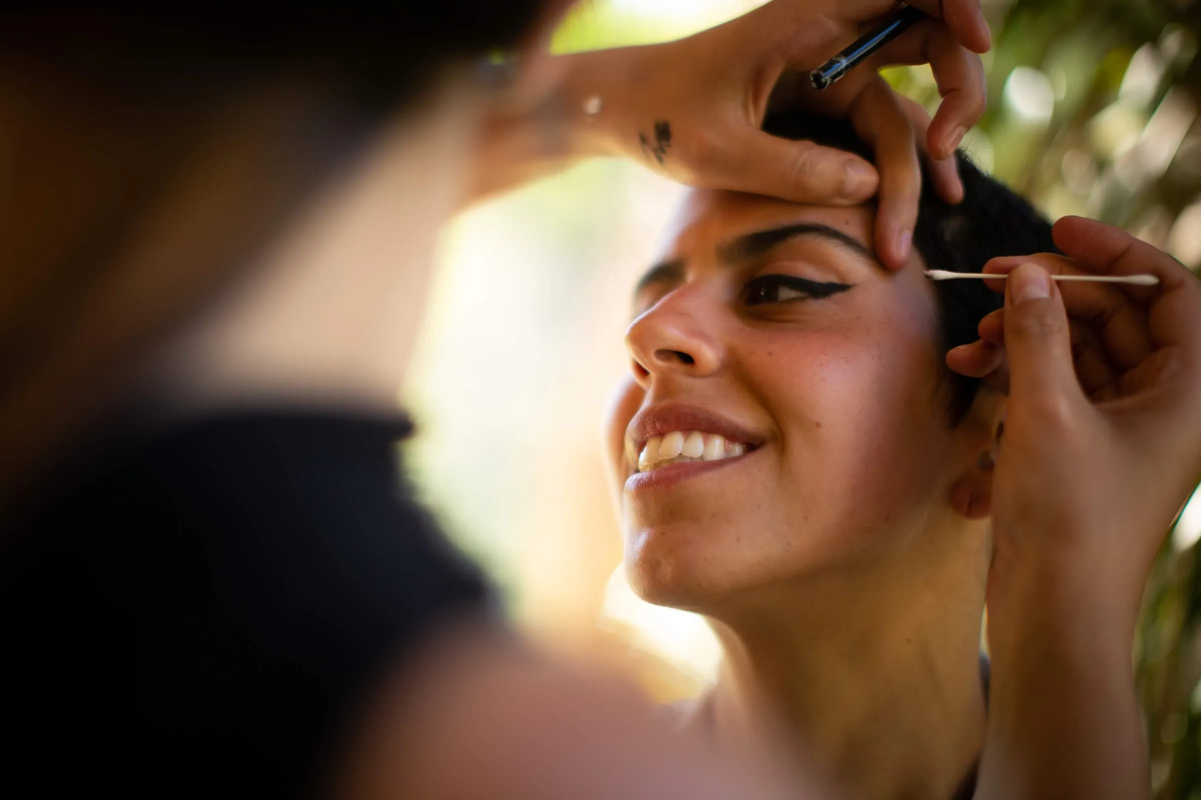 Close da noiva sorrindo durante a maquiagem, em contraluz dourado, na Fazenda Cósmica