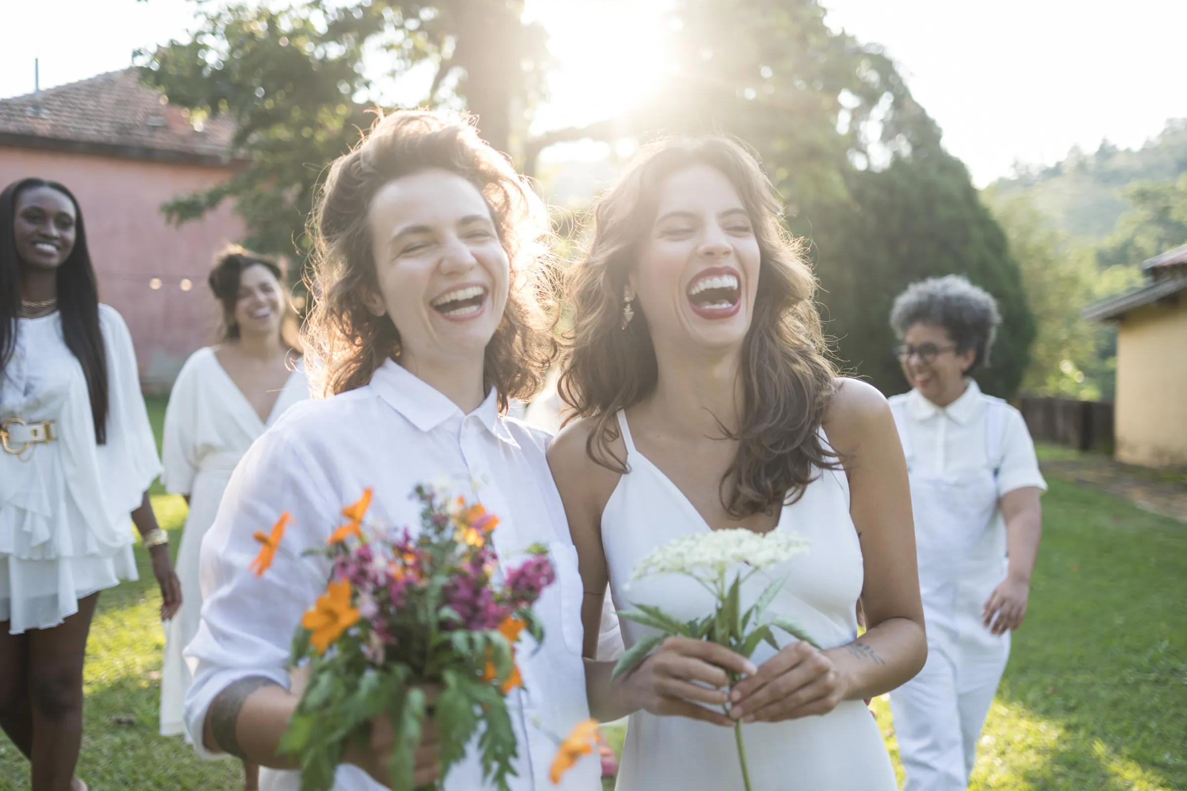 Duas noivas rindo em contraluz de fim de tarde, com buquês de flor do campo, na Fazenda Cósmica