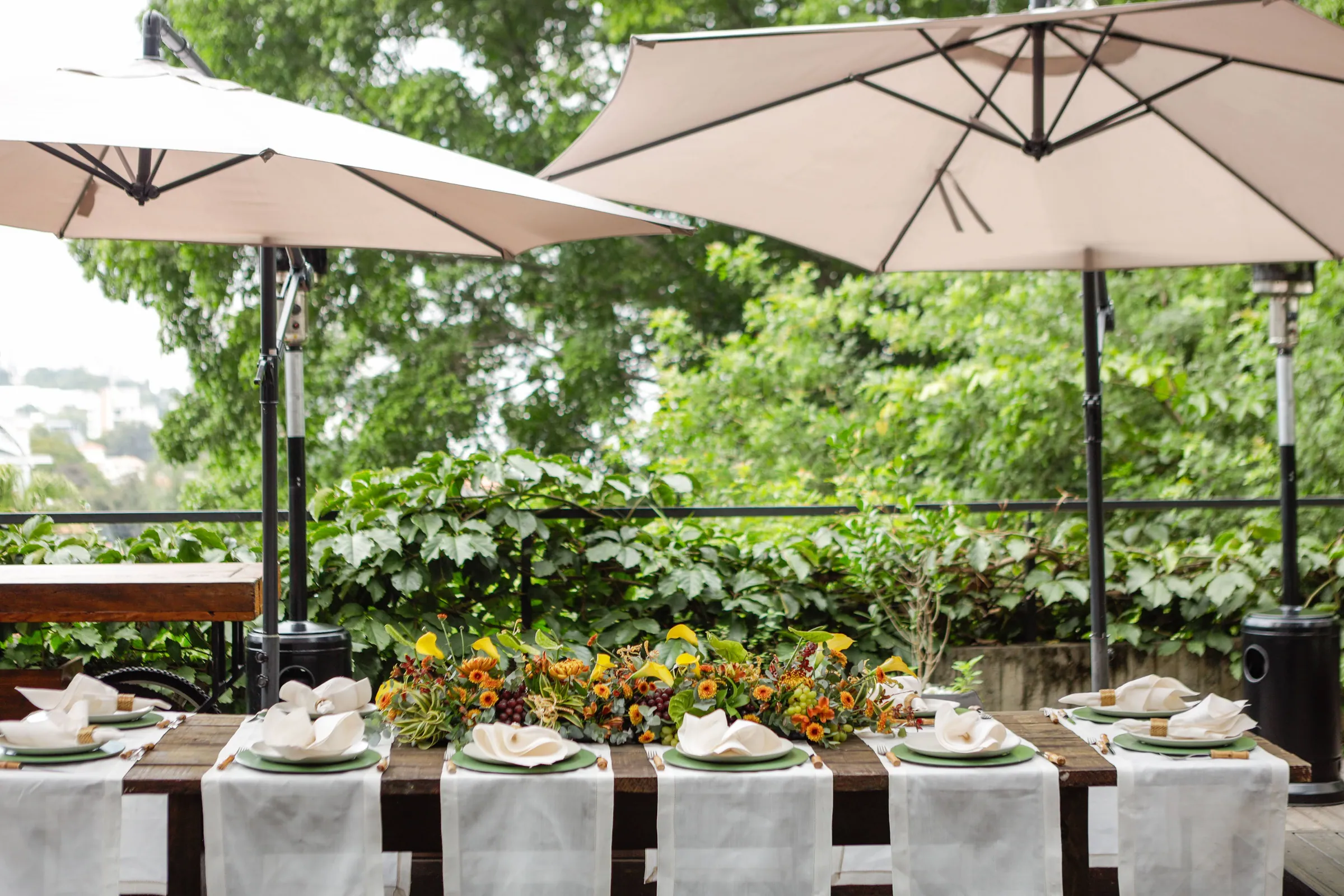 Mesa posta para o casamento embaixo de guarda-sóis bege, com arranjo floral em tons de amarelo e vinho ao centro e a vegetação do jardim ao fundo