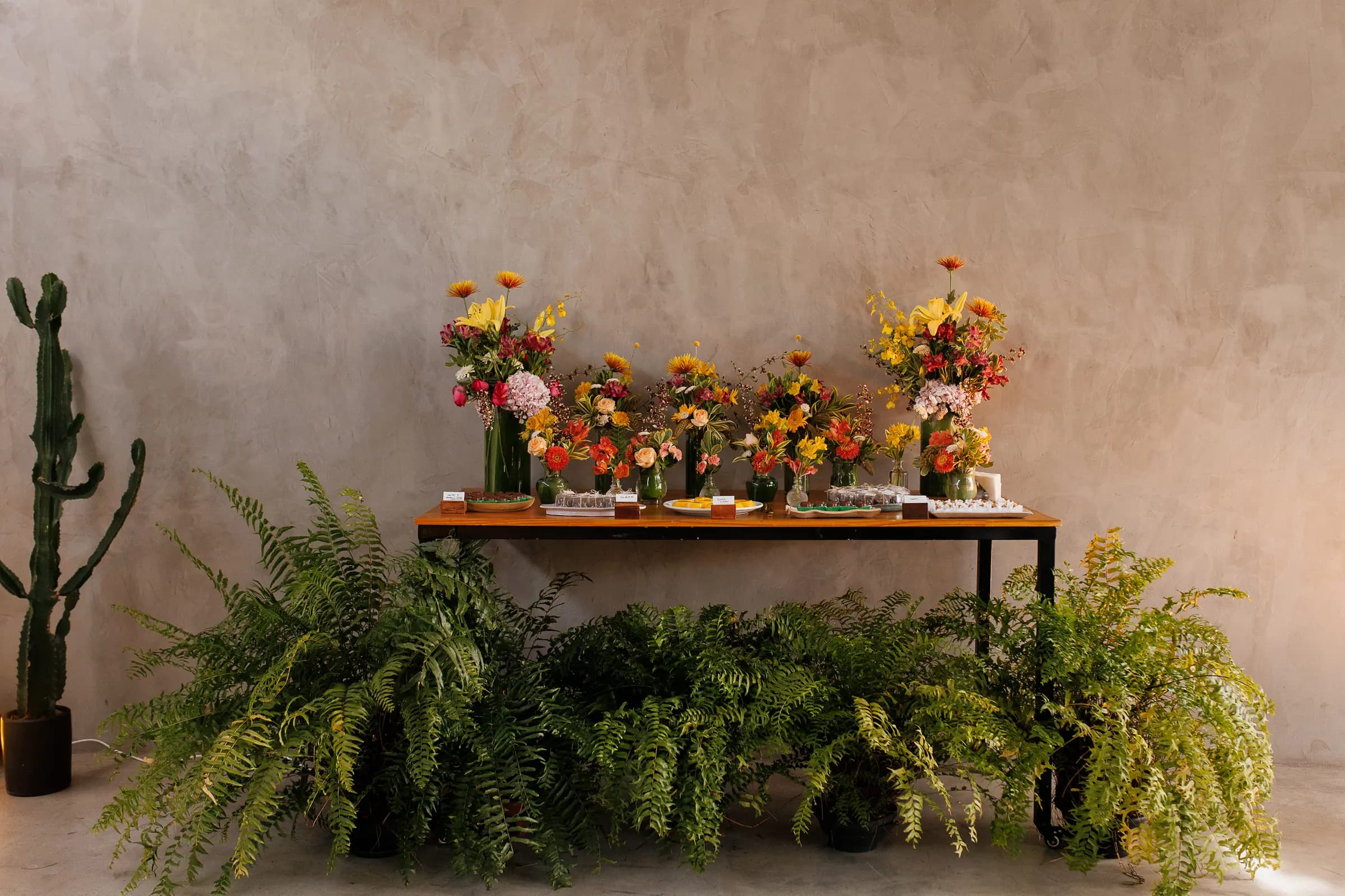 Mesa de flores do casamento, com vasinhos variados em tons de amarelo e vermelho contra a parede de textura rústica, cacto e samambaias ao redor