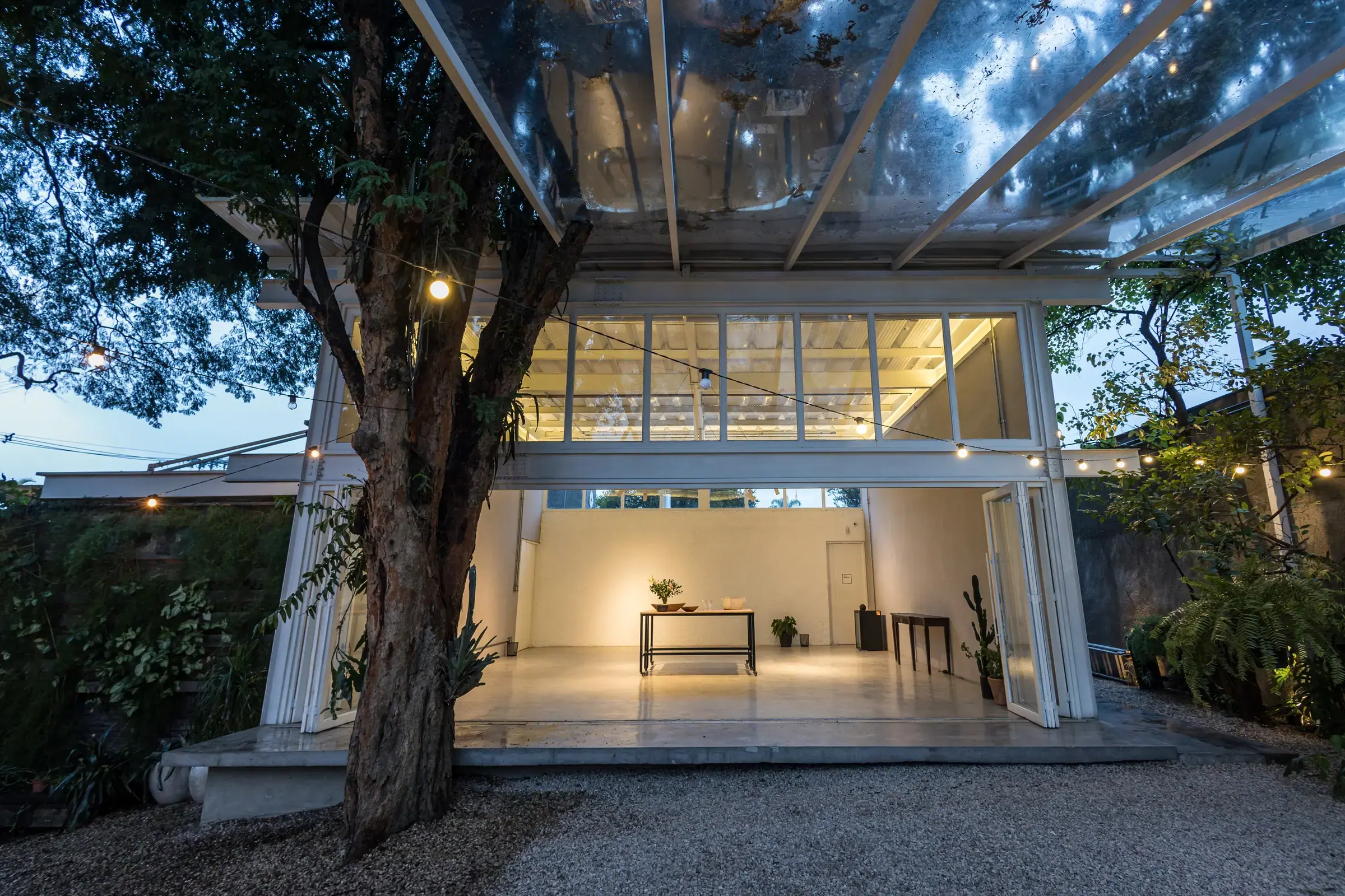 Fachada do espaço de eventos ao entardecer, com luzes de barbante entre as árvores e o salão iluminado ao fundo, pronto para receber o casamento