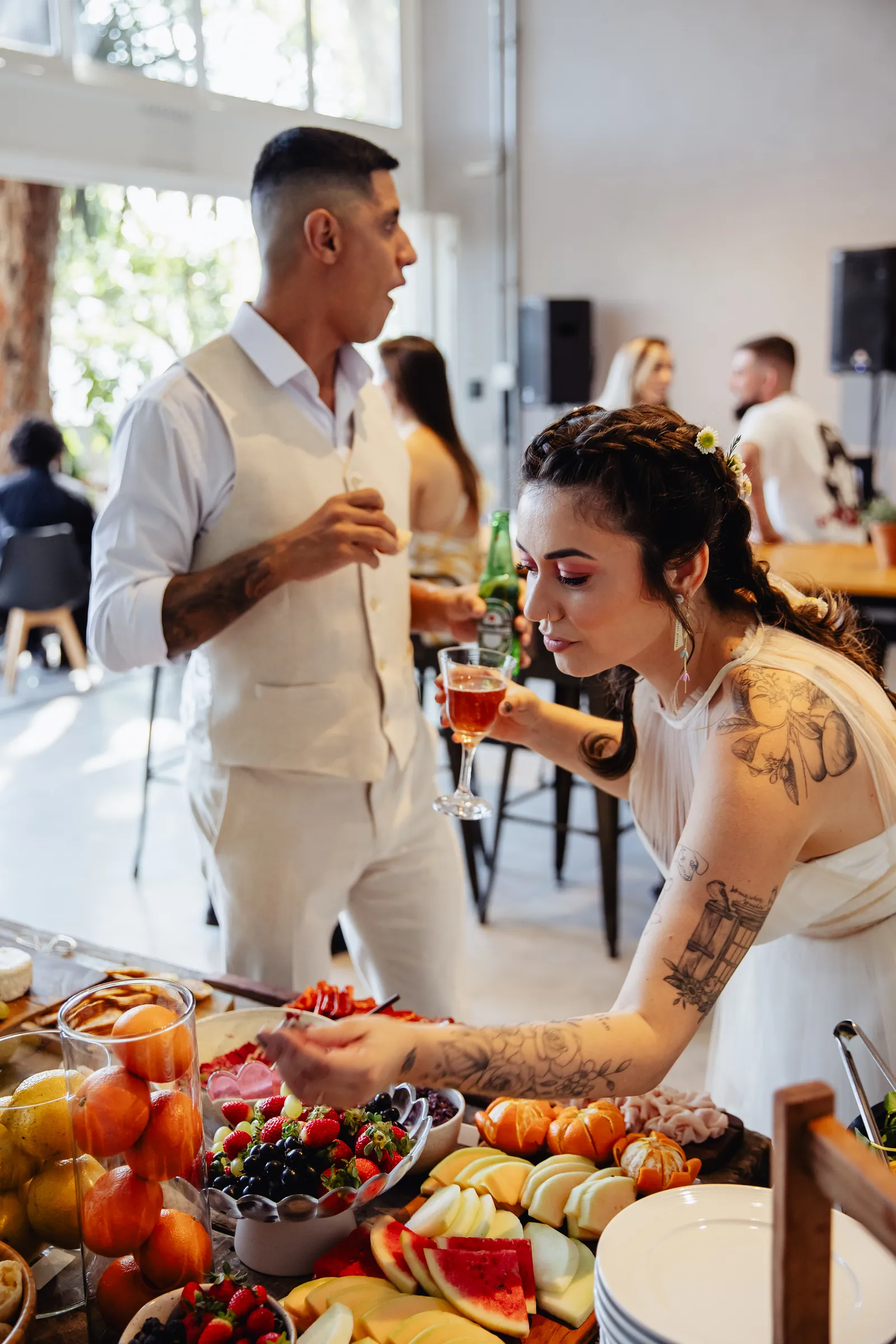 Noivos conversando à mesa de frutas e frios durante a festa de casamento, ela brindando com uma taça de vinho