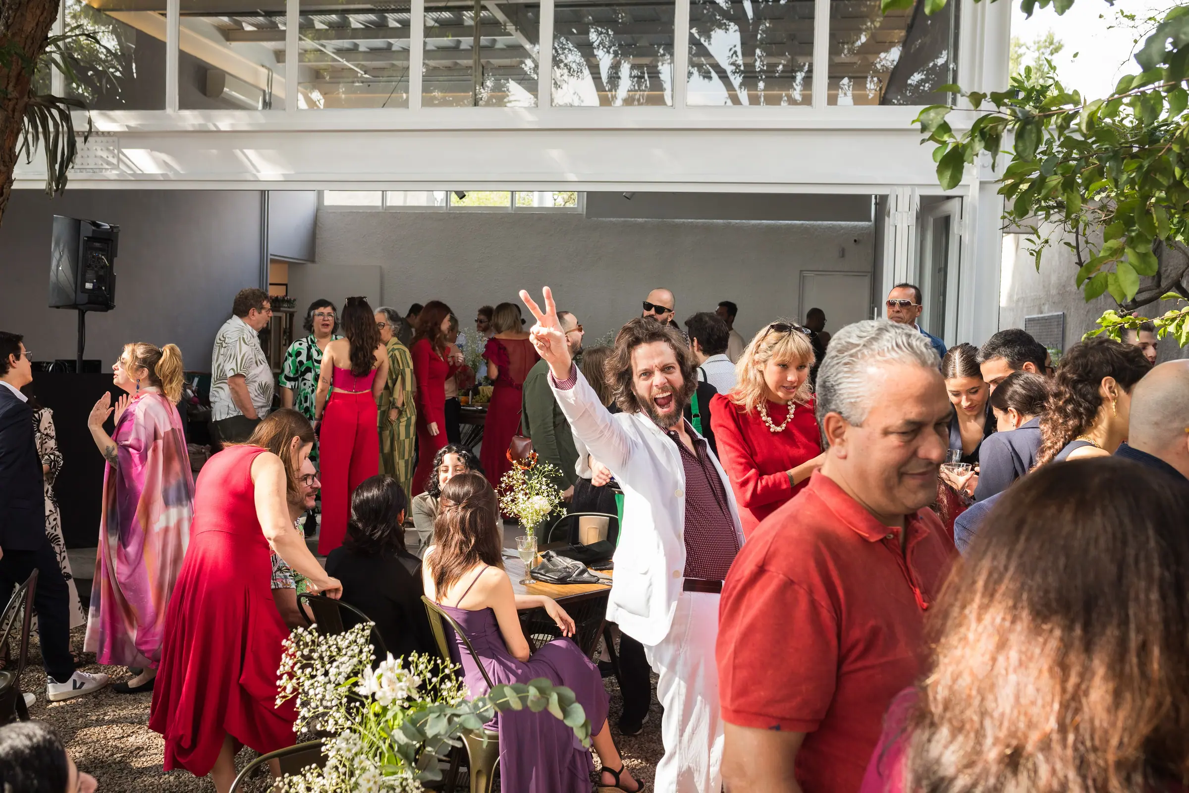 Festa de casamento animada, convidado erguendo os dedos em V e sorrindo em meio a convidados vestidos de vermelho e rosa