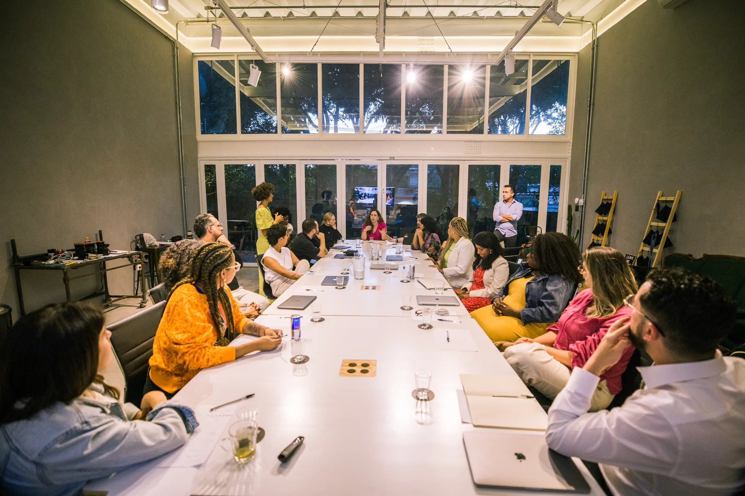 Workshop em formato plenária na Casa Cósmica, mesa única comprida iluminada à noite com cerca de quinze pessoas em volta, pé-direito alto e janelões