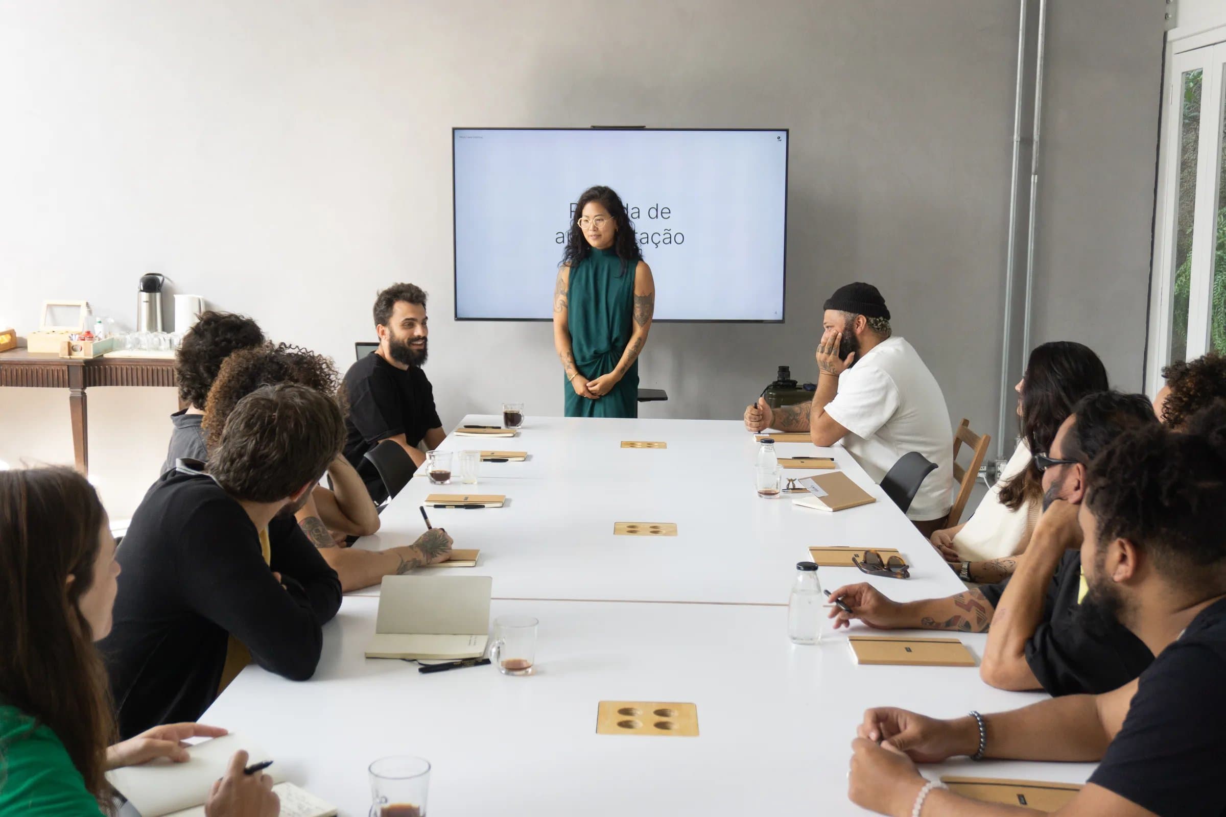 Apresentação em reunião na Casa Cósmica, facilitadora de vestido verde em pé diante da TV em frente a grupo sentado em mesa branca, sala minimalista