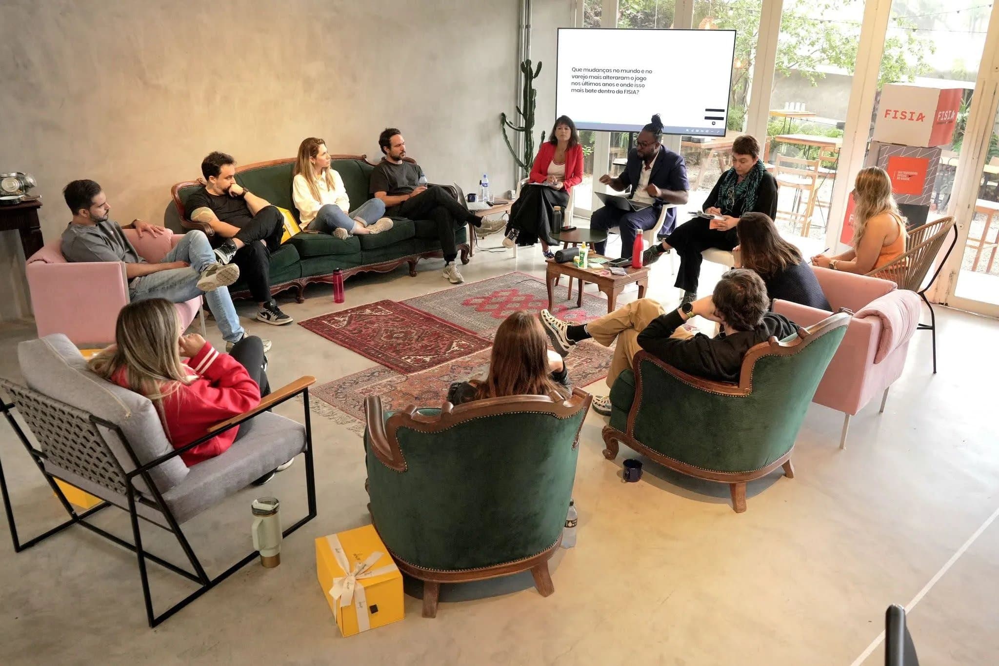 Reunião corporativa em formato lounge na Casa Cósmica, equipe sentada em sofás verdes de veludo e poltronas rosa em torno de tapete persa, telão atrás
