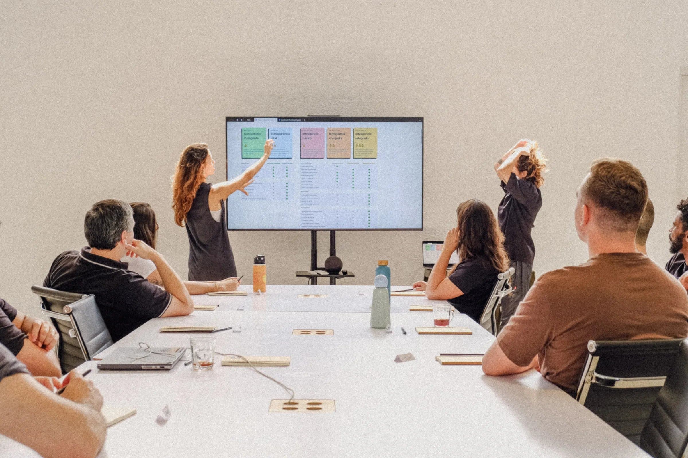 Revisão de sprint na Casa Cósmica, facilitadora em pé apontando para painel de post-its coloridos na TV diante de equipe sentada em mesa branca
