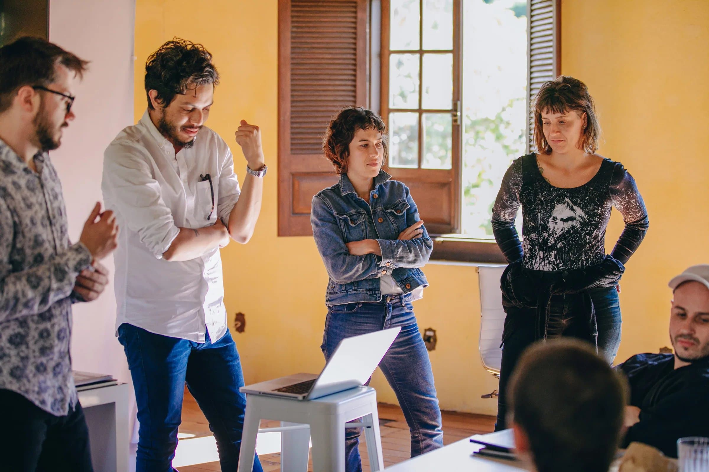 Briefing em pé na sala amarela da Fazenda Cósmica durante treinamento, grupo em volta de notebook combinando próximos passos