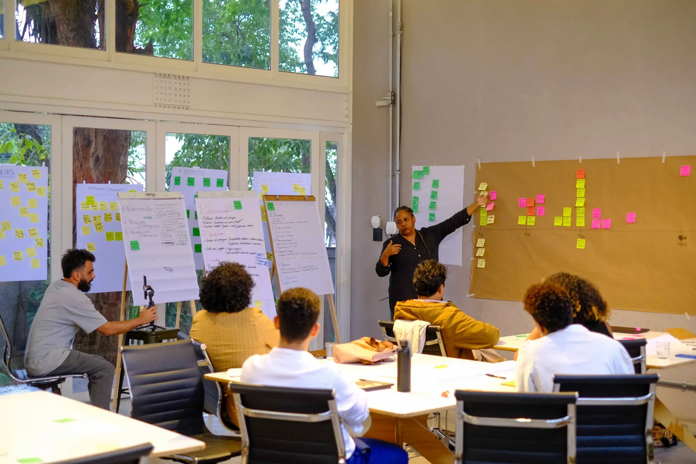 Workshop corporativo na Casa Cósmica, em São Paulo: sala modular com mesas em ilhas, facilitador apresentando frente a painel de post-its, equipe sentada em volta com janelão para o jardim.