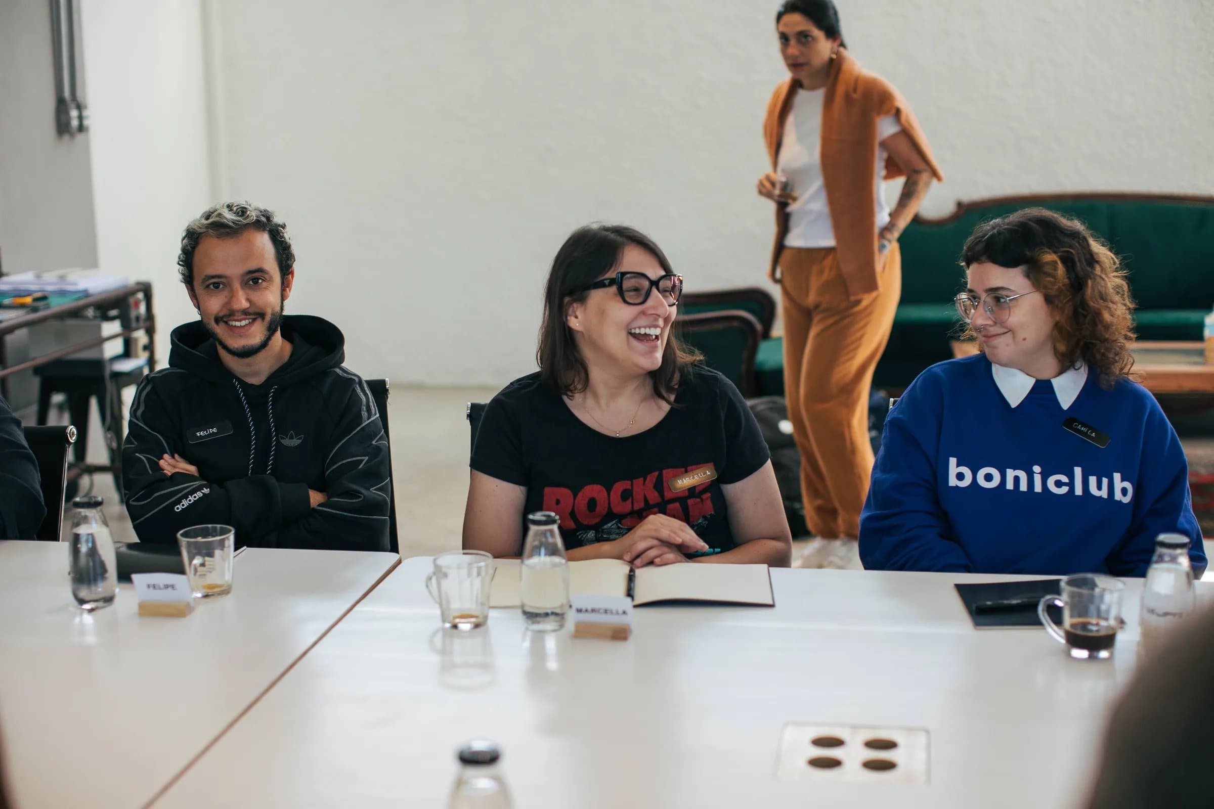 Workshop criativo na Casa Cósmica: três colegas de equipe de marketing rindo durante a dinâmica em torno da mesa, clima leve e descontraído.