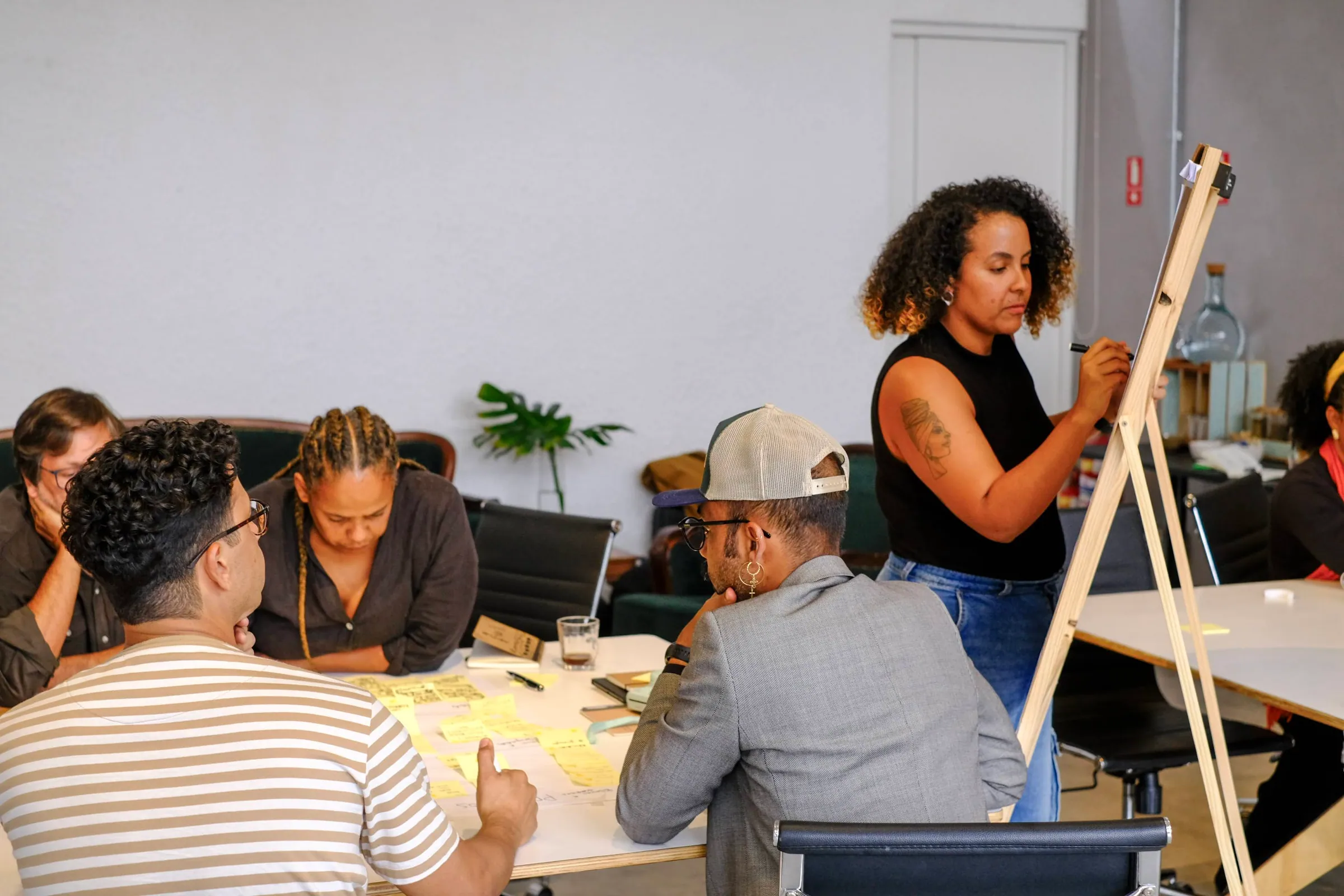 Equipe reunida ao redor de mesa com post-its coloridos, enquanto uma integrante desenha num cavalete de madeira