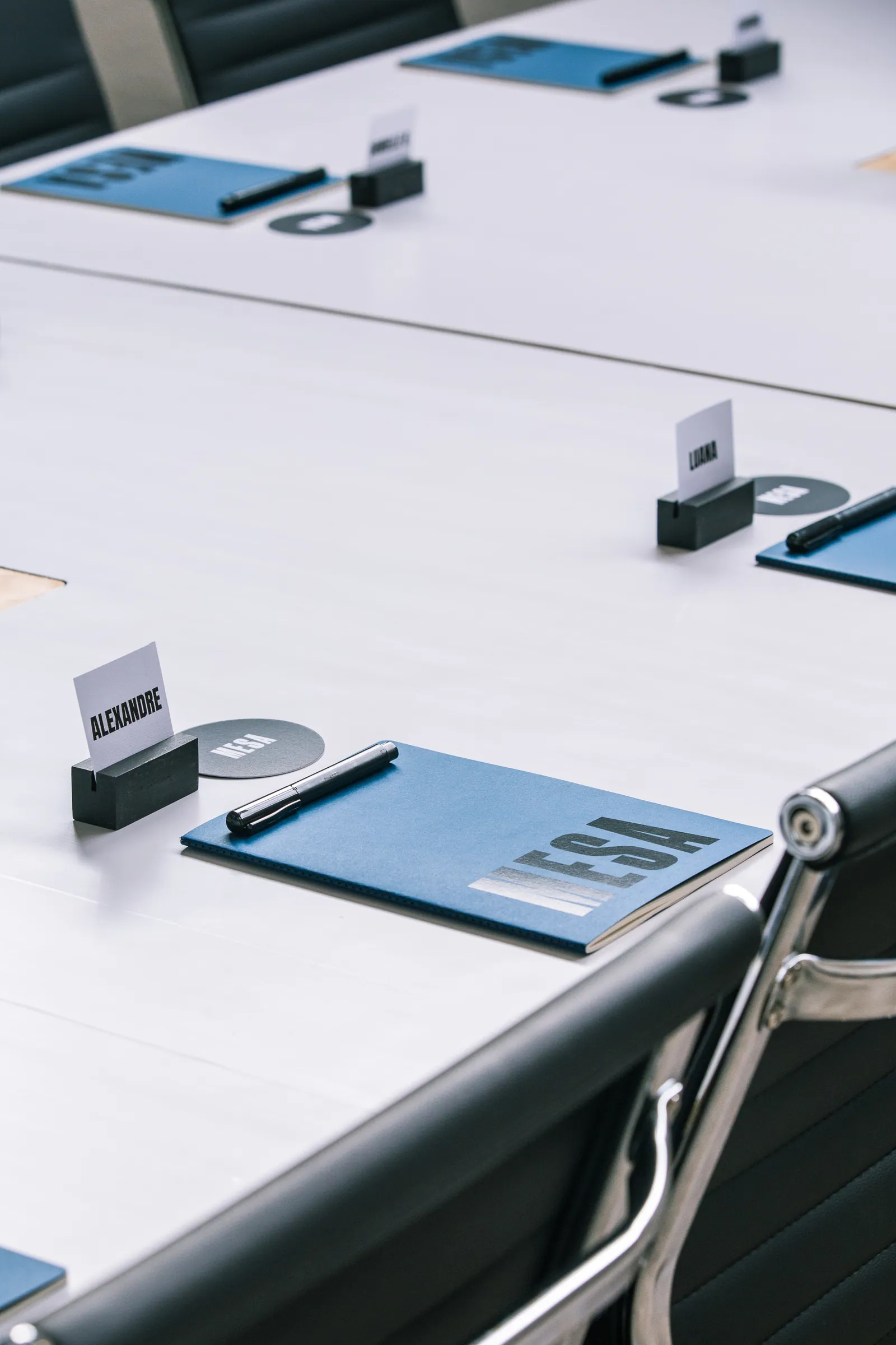 Detalhe de mesa de reunião com caderno azul personalizado, caneta e crachá de identificação sobre a mesa