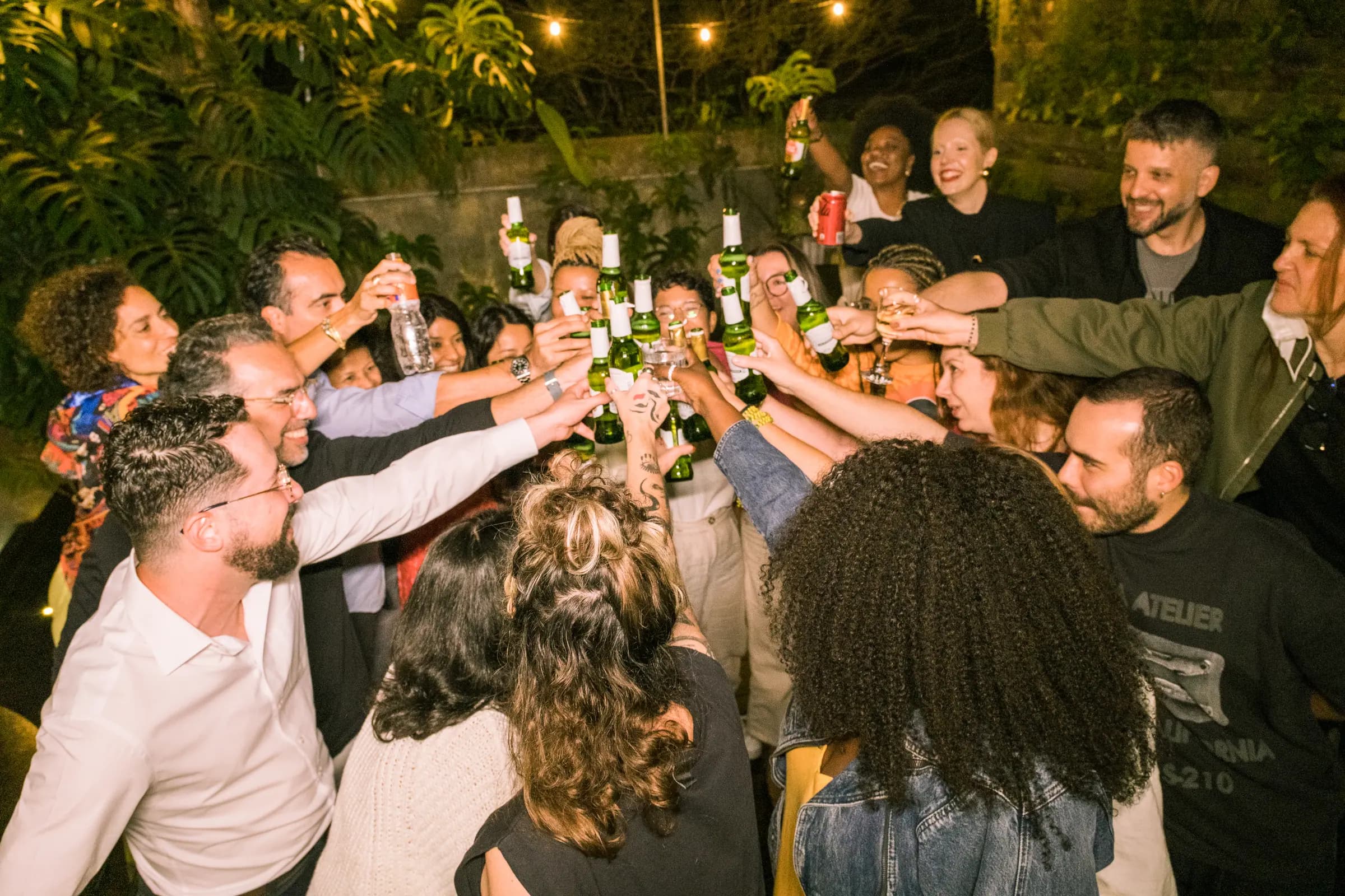 Grupo grande brinda com garrafas de cerveja erguidas numa confraternização noturna no jardim, sob luzes penduradas