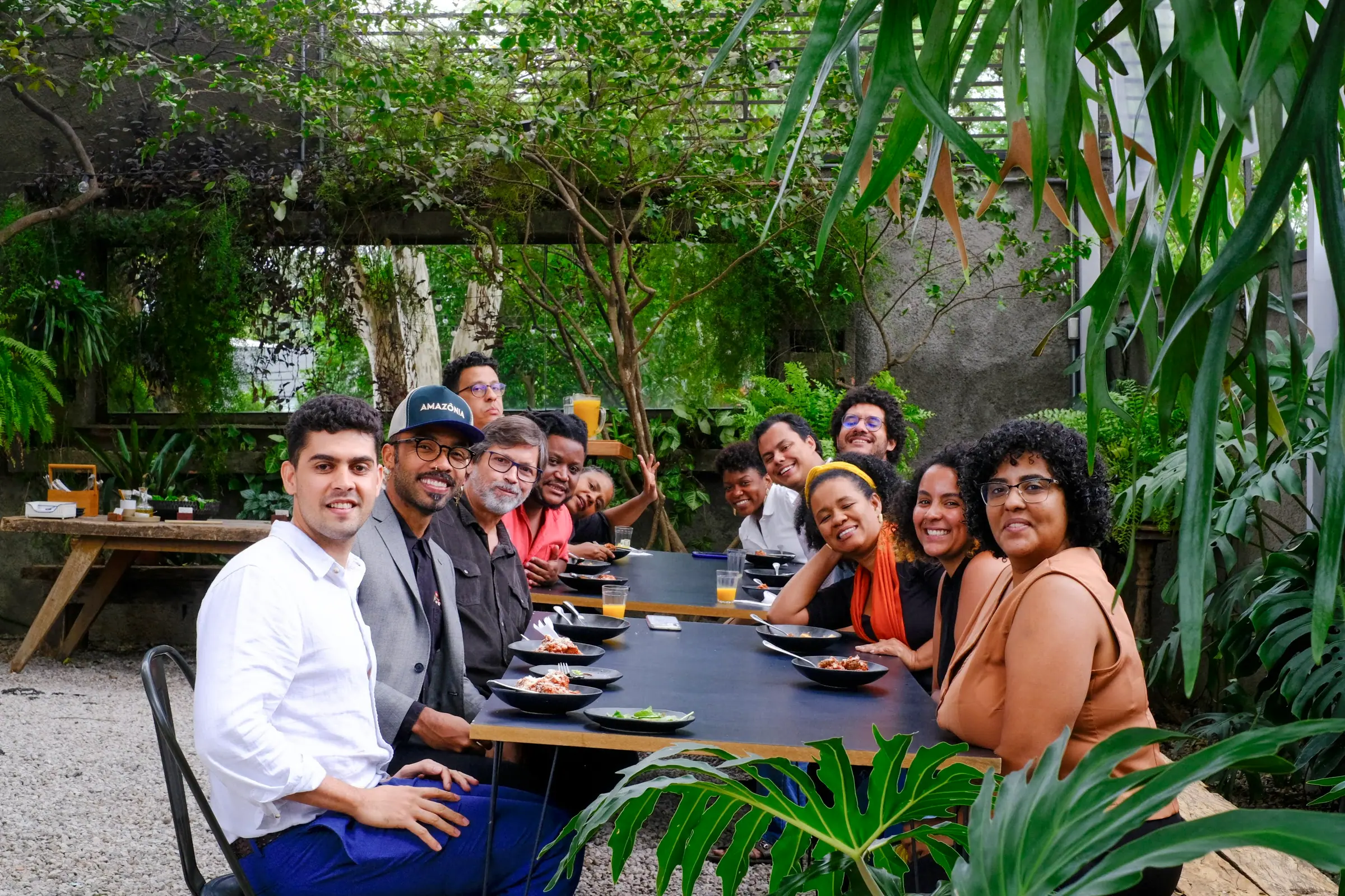 Grupo posa sorrindo à mesa durante almoço sob pergolado verde na Casa Cósmica, pratos servidos à frente