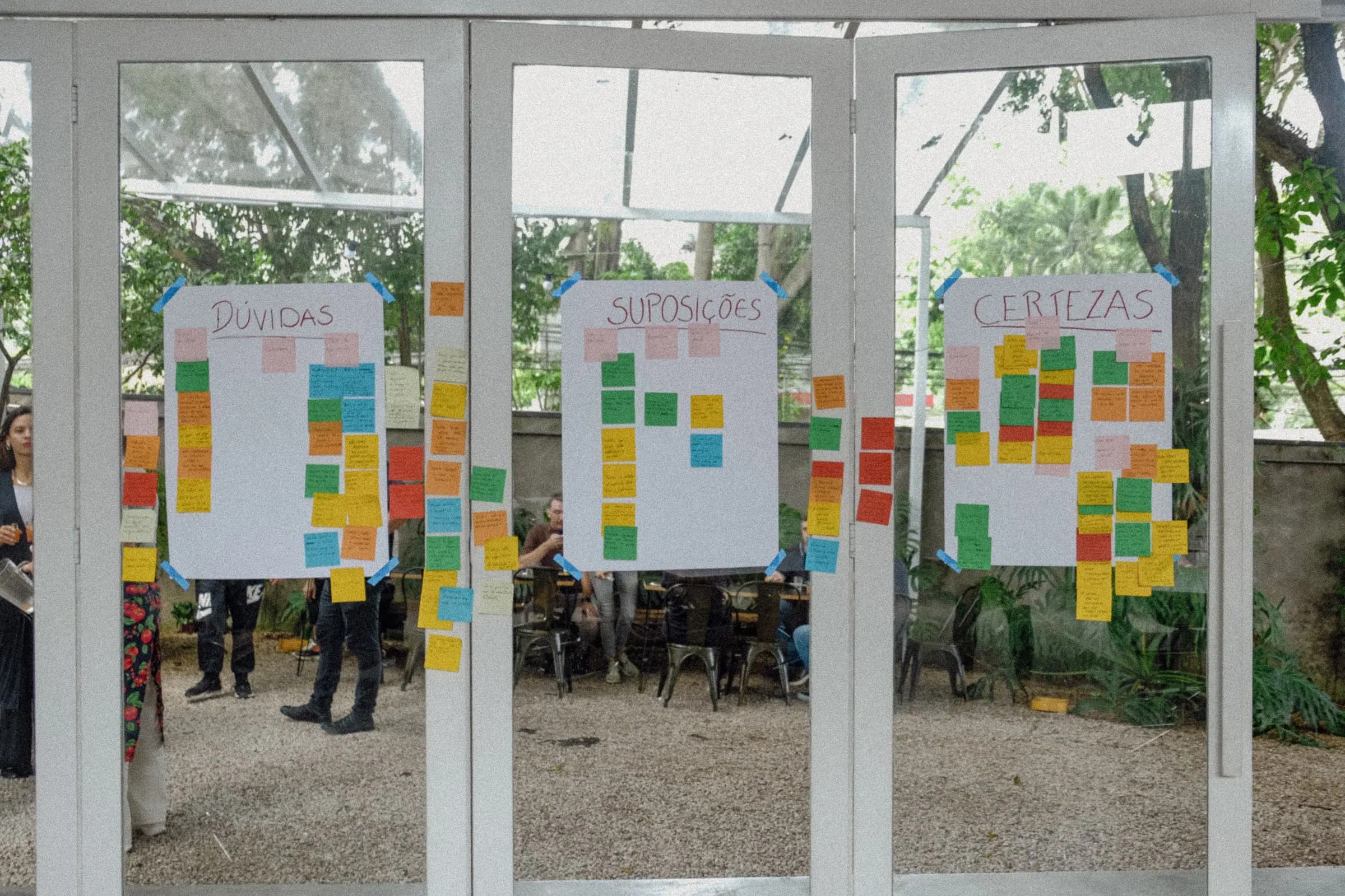 Painéis com post-its coloridos organizados em 'Dúvidas', 'Suposições' e 'Certezas', vistos através das portas de vidro da Casa Cósmica