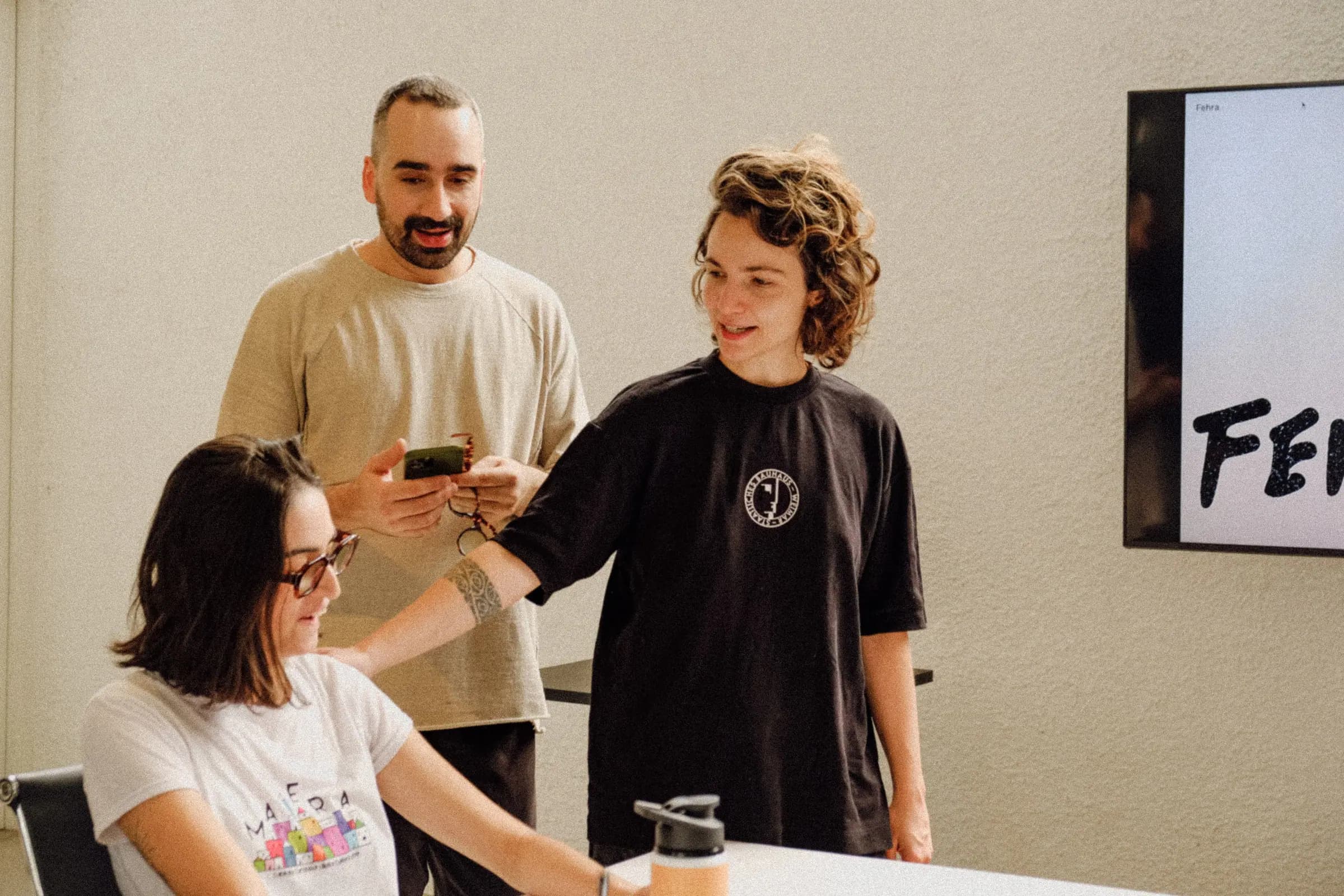 Colegas de equipe sorriem enquanto olham um celular, tela ao fundo com o texto 'FEHRA'