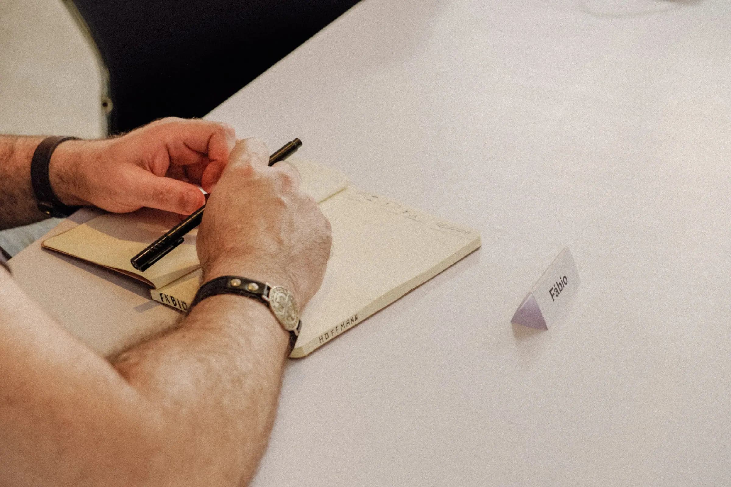 Mão escrevendo em caderno de anotações ao lado de crachá de mesa com o nome 'Fábio'