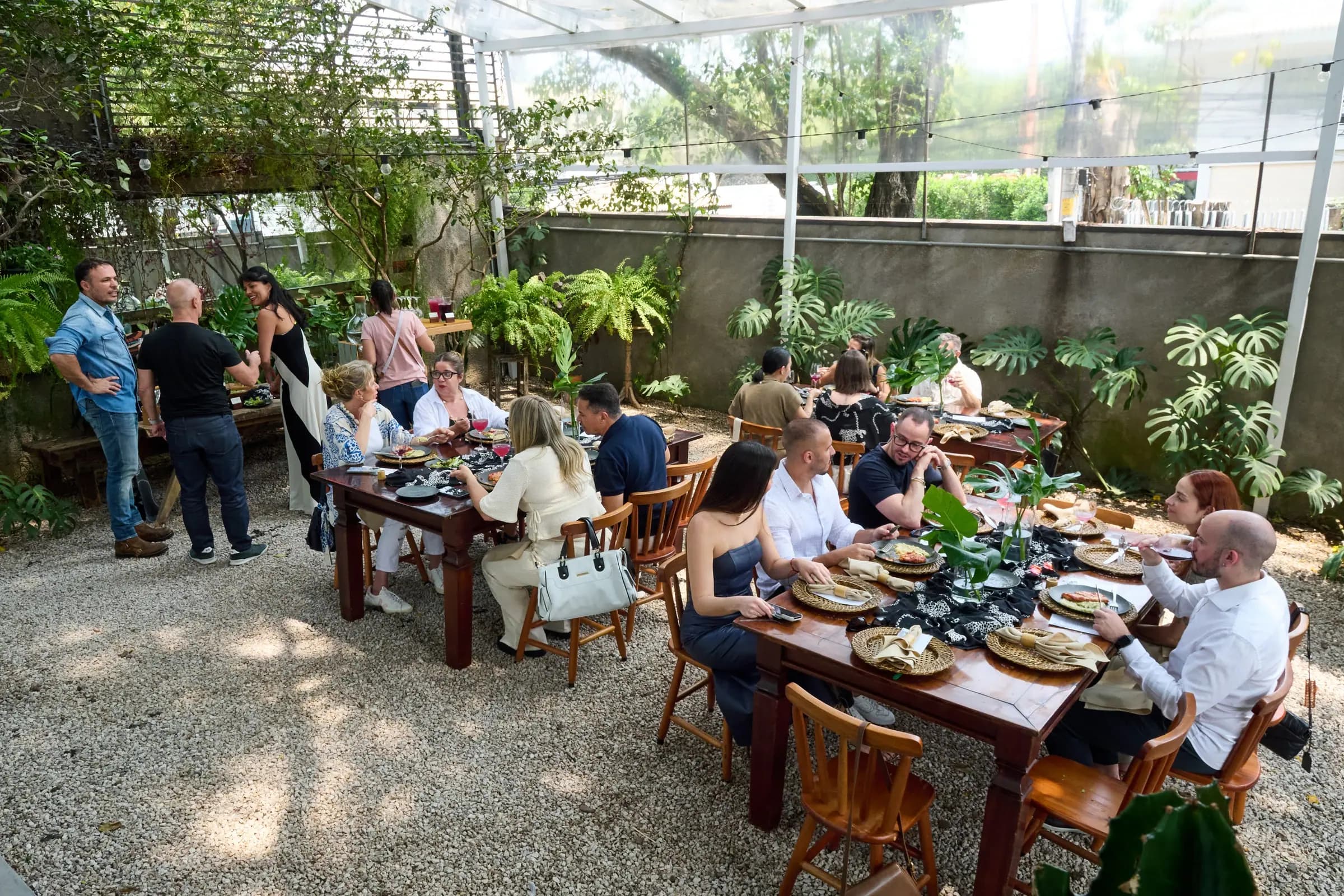 Convidados almoçam em mesas dispostas no jardim coberto da Casa Cósmica, luz filtrada pela vegetação