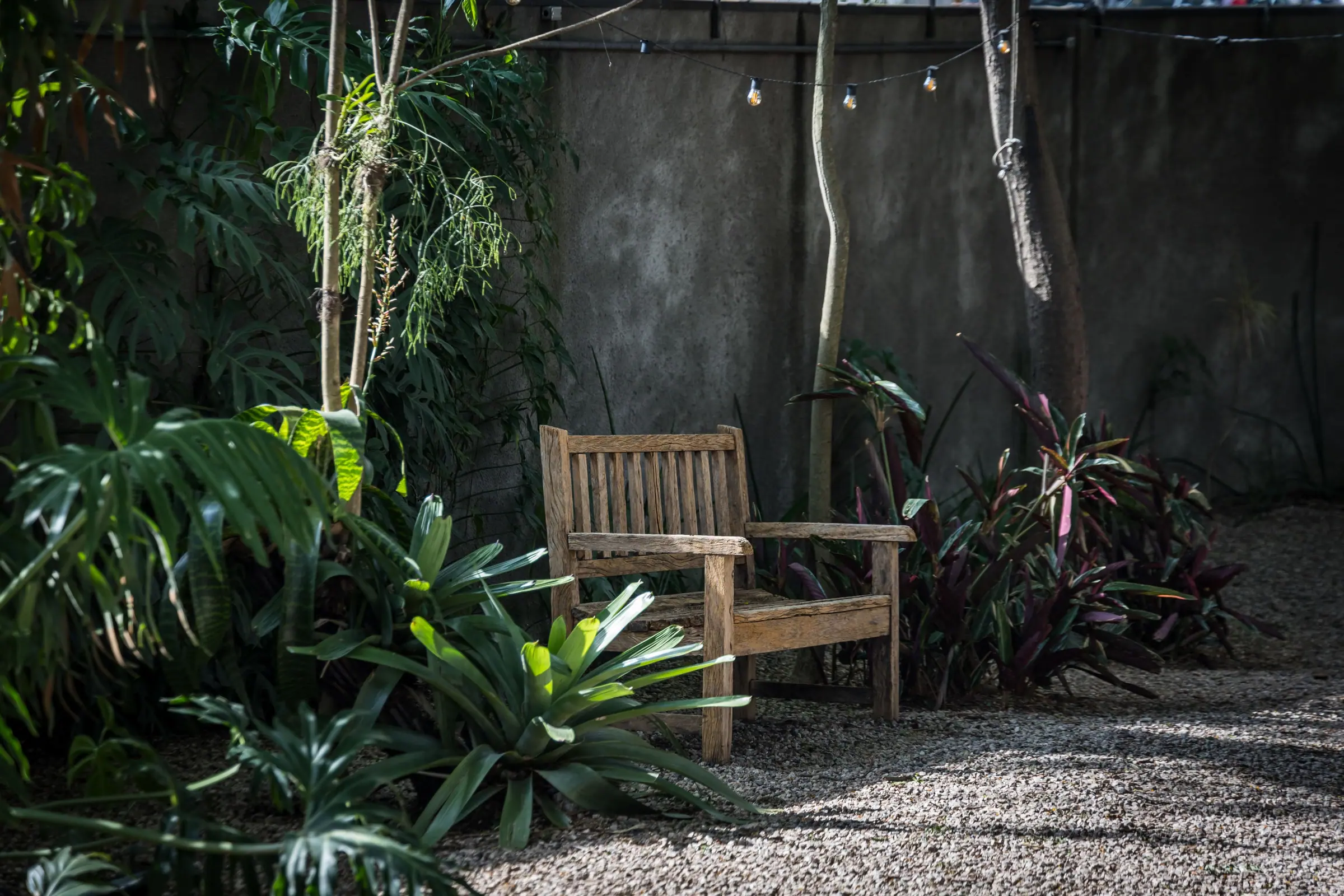 Cadeiras de metal enfileiradas junto a mesa de madeira no jardim de pedrisco da Casa Cósmica, luzes de fio acesas entre as árvores