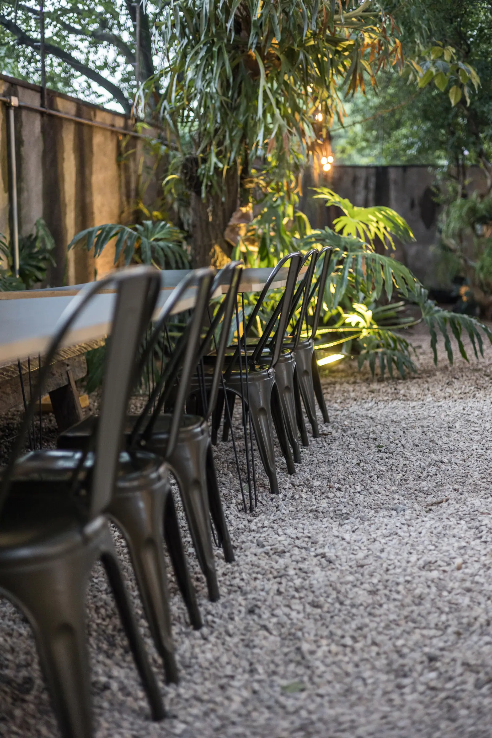 Mesa comprida com cadeiras de metal enfileiradas no jardim de pedrisco, luzes de fio acesas ao entardecer na Casa Cósmica