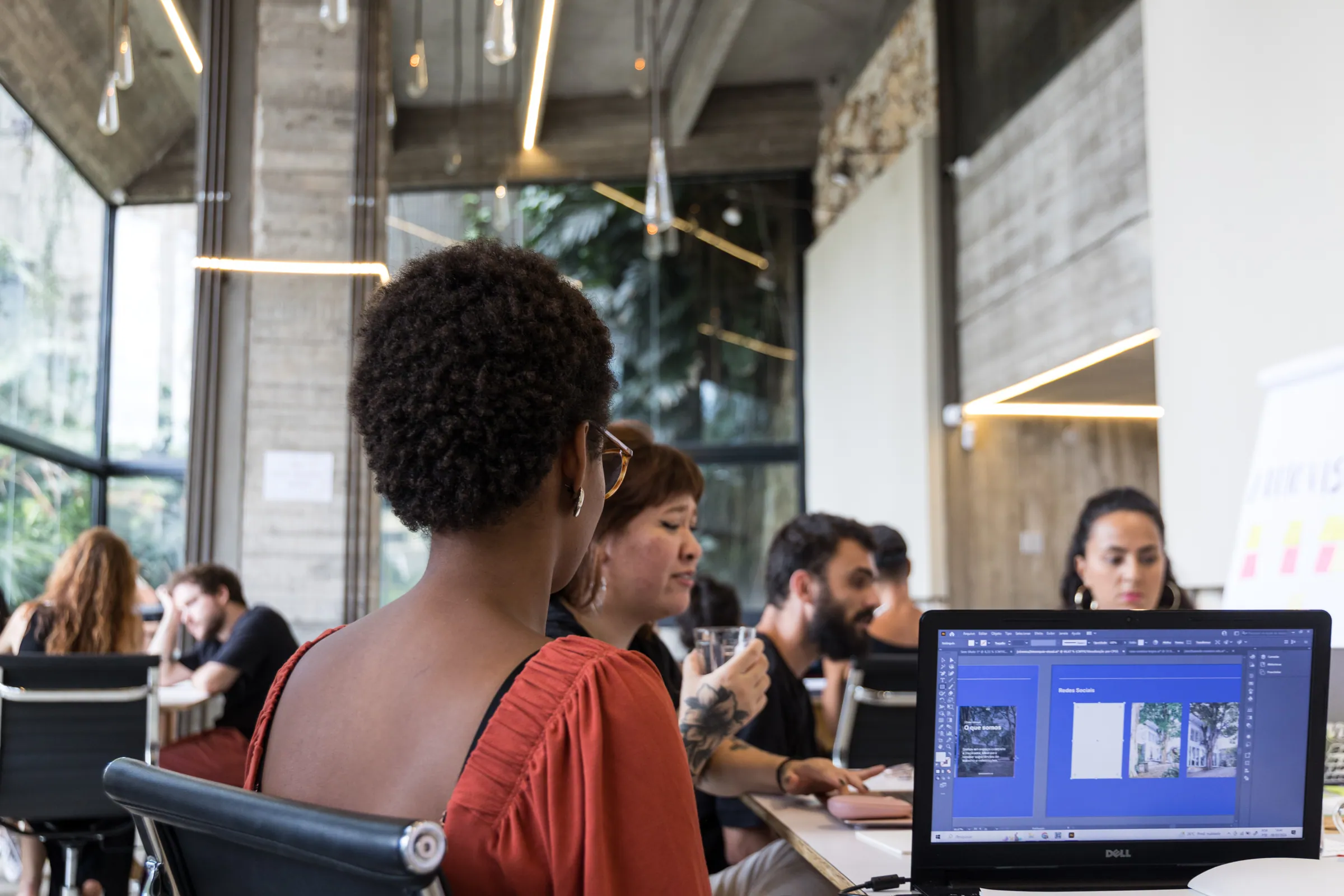 Profissional trabalha em notebook com apresentação de 'Redes Sociais' na tela, colegas ao fundo em ambiente com luminárias suspensas