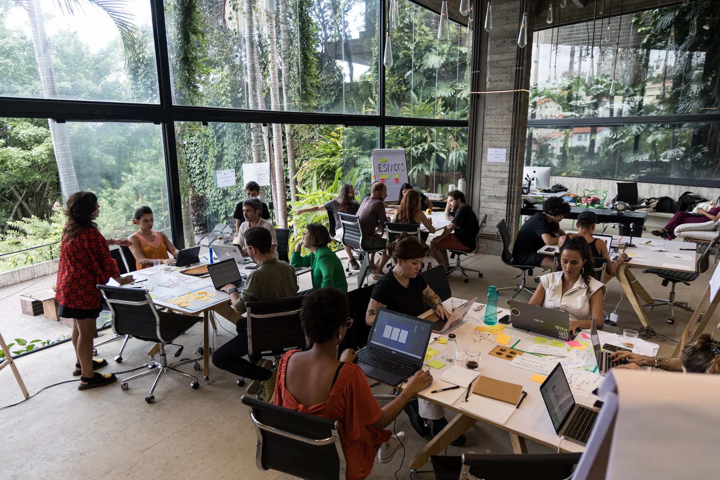 Equipe distribuída em mesas com notebooks abertos, cartaz 'Espaços' fixado no vidro ao fundo, vista para o jardim