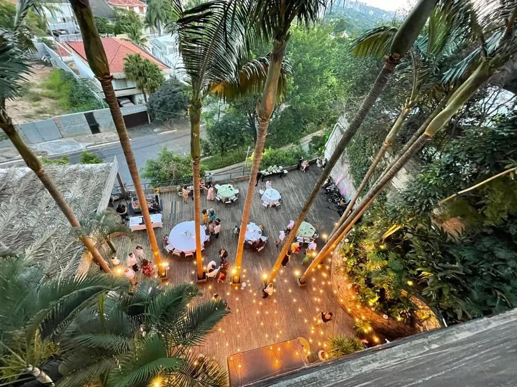 Vista aérea de terraço decorado para evento ao entardecer, mesas redondas entre palmeiras e luzes no chão