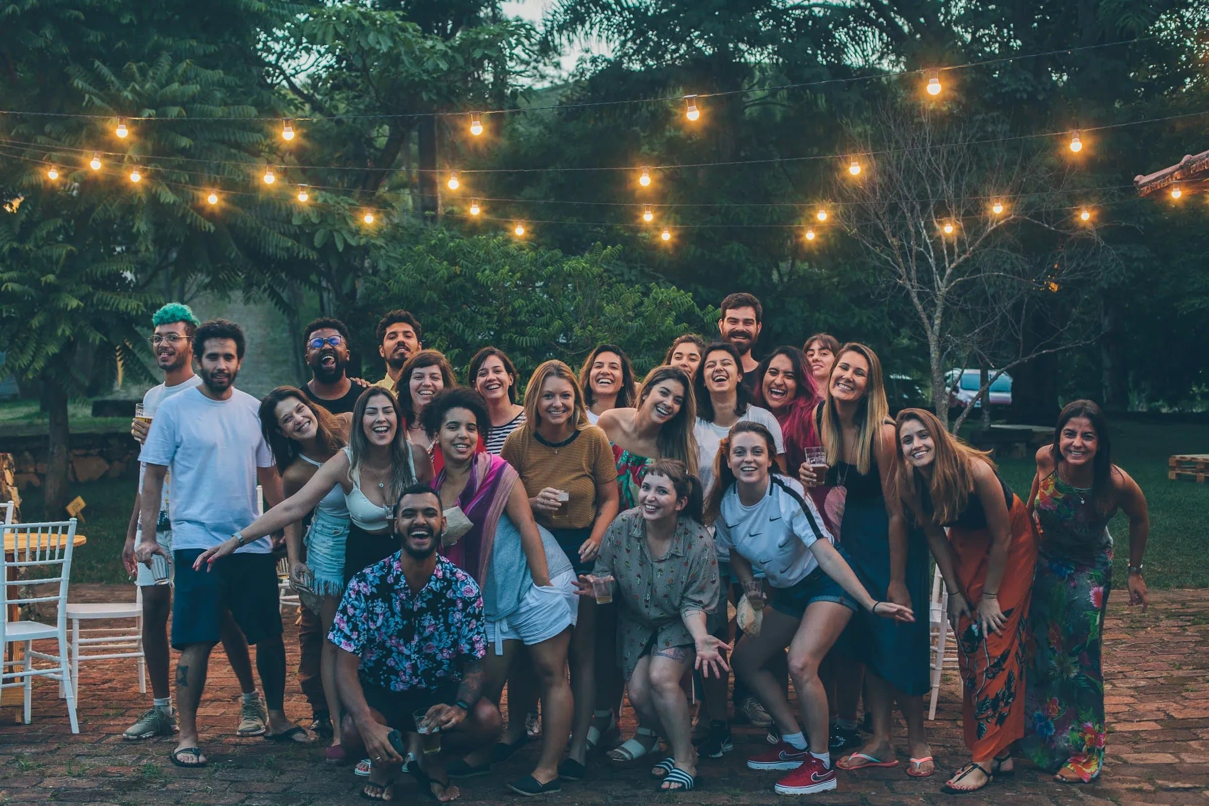 Foto de grupo grande em confraternização na Fazenda Cósmica, cerca de vinte e cinco pessoas sorrindo juntas sob varal de luzes amarelas ao entardecer
