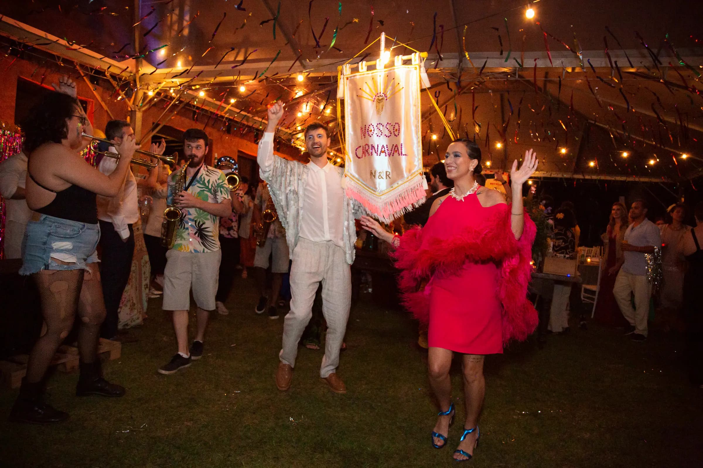 Bloco de carnaval com fanfarra e convidados fantasiados, sob luzes penduradas